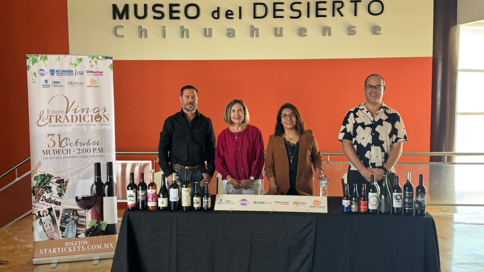 Cuatro personas posan detrás de una mesa con botellas de vino junto a un cartel del festival en el Museo del Desierto Chihuahuense.